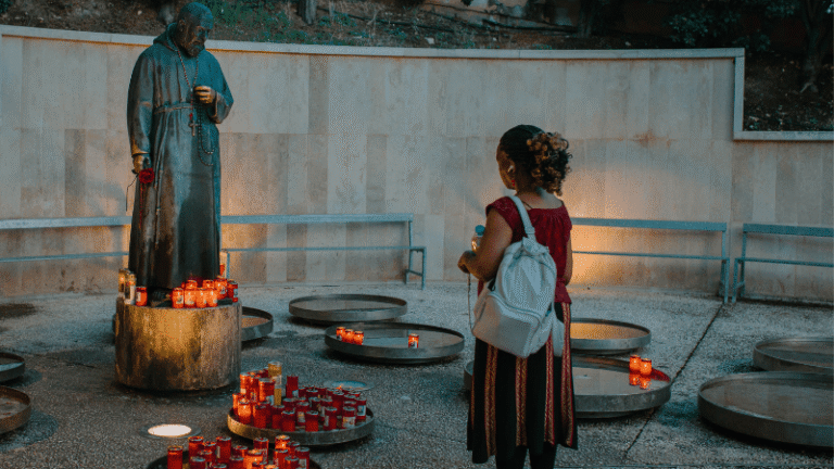 Pellegrina in preghiera davanti alla statua di Padre Pio circondata da candele votive accese, nel piazzale esterno del santuario di San Giovanni Rotondo.