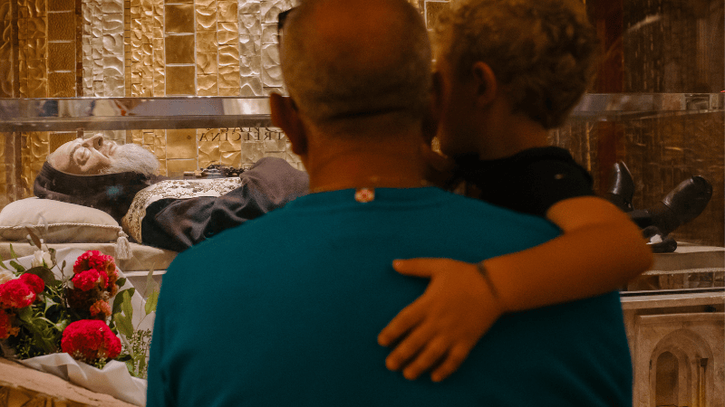 Un uomo con un bambino in braccio prega davanti al corpo di Padre Pio esposto nella cripta del santuario di San Giovanni Rotondo.