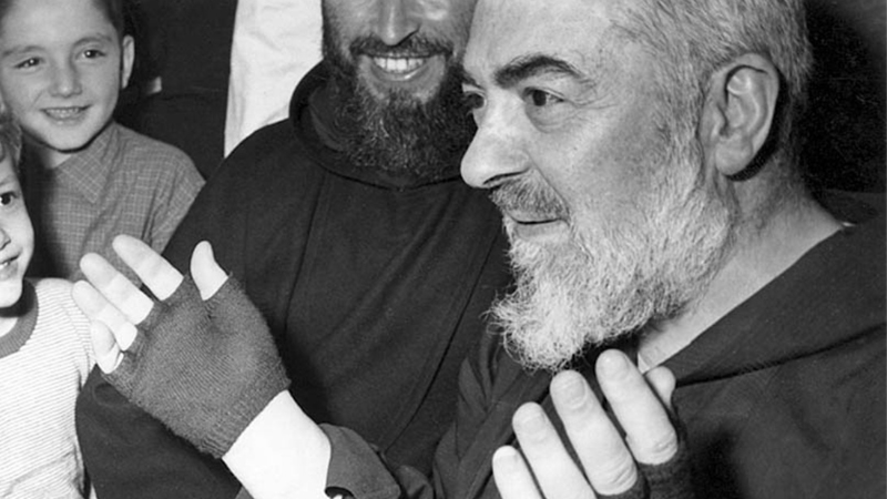 Padre Pio da Pietrelcina mostra le mani coperte dai guanti per le stimmate, sorridendo tra i fedeli in un momento di incontro e devozione.