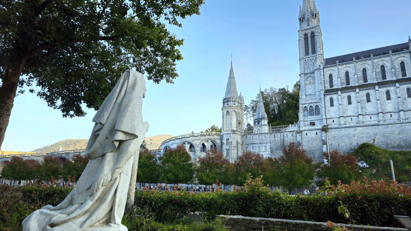 Statua di Bernadette Soubirous in preghiera rivolta verso la maestosa Basilica di Lourdes, in un'atmosfera di pace e devozione.
