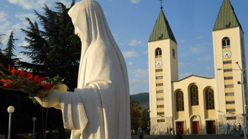 Statua della Vergine Maria con fiori davanti alla celebre Chiesa di San Giacomo, cuore spirituale del Santuario di Medjugorje.