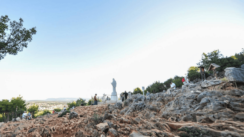Ampia veduta del Podbrdo a Medjugorje con pellegrini in preghiera presso la statua della Madonna, luogo delle apparizioni mariane.