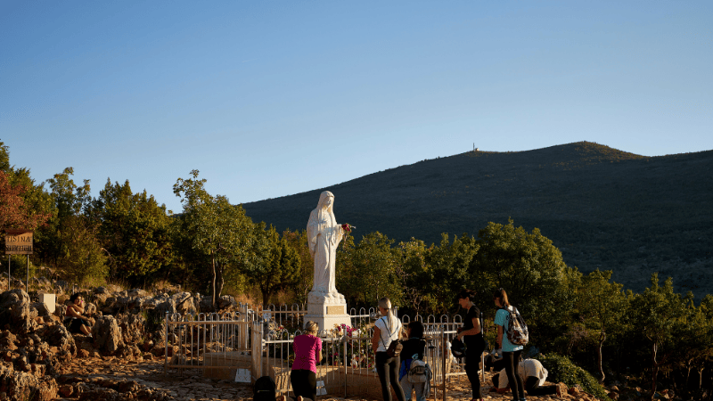 Fedeli raccolti intorno alla statua della Vergine Maria sul Podbrdo a Medjugorje, luogo di speranza e preghiera per molte famiglie.