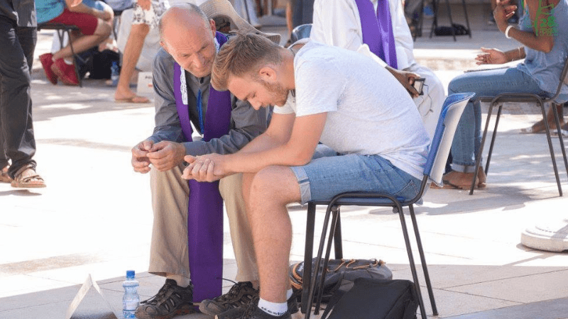 Giovane si confessa con sacerdote in stola viola durante pellegrinaggio a Medjugorje all'aperto con altri pellegrini